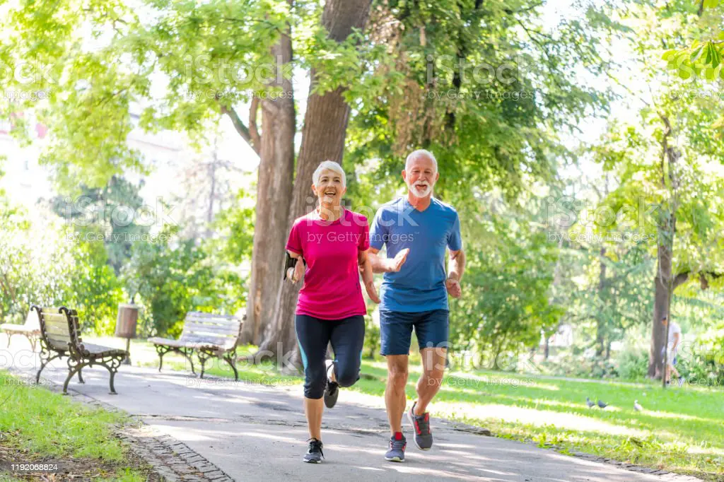 Mature Sporty Couple Jogging Outdoors in Nature. Two Elderly Athlete Persons is Running in the Park on a Sport Track During a Warm Sunny Day.